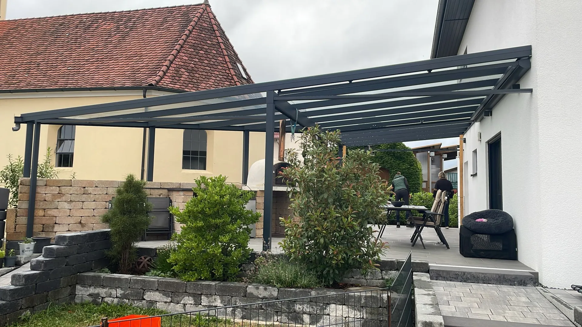 Terrassenüberdachung mit dunklen Profilen über einer Terrasse mit Pflanzen und Sitzplatz.