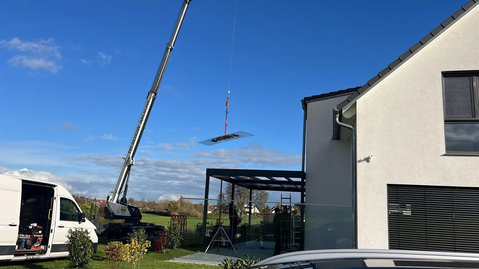 Ein Kran hebt ein Dachelement über eine Terrassenüberdachung neben einem Wohnhaus.