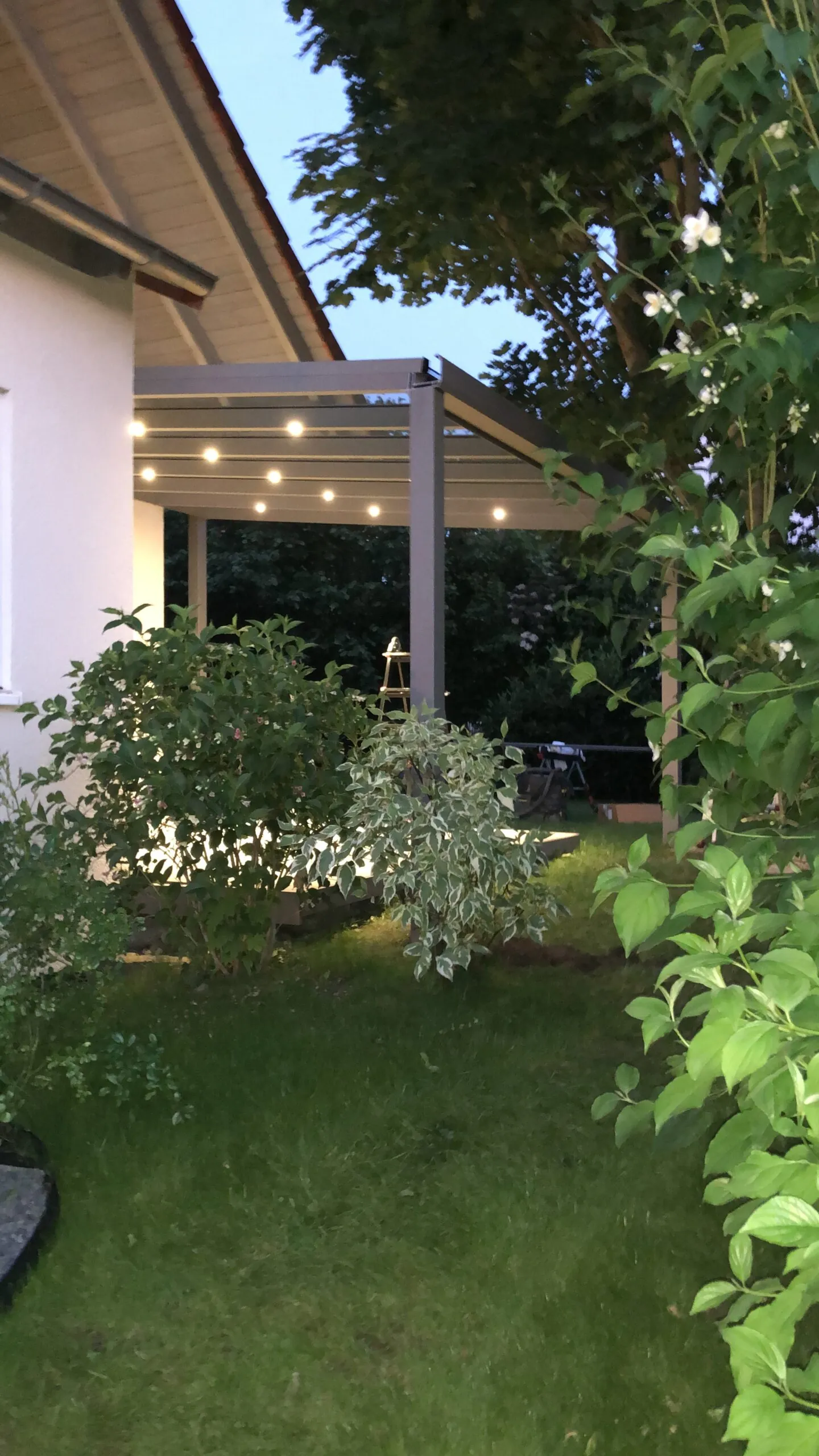 Blick aus dem bepflanzten Garten auf eine Terrassenüberdachung mit eingeschalteten Dachleuchten.
