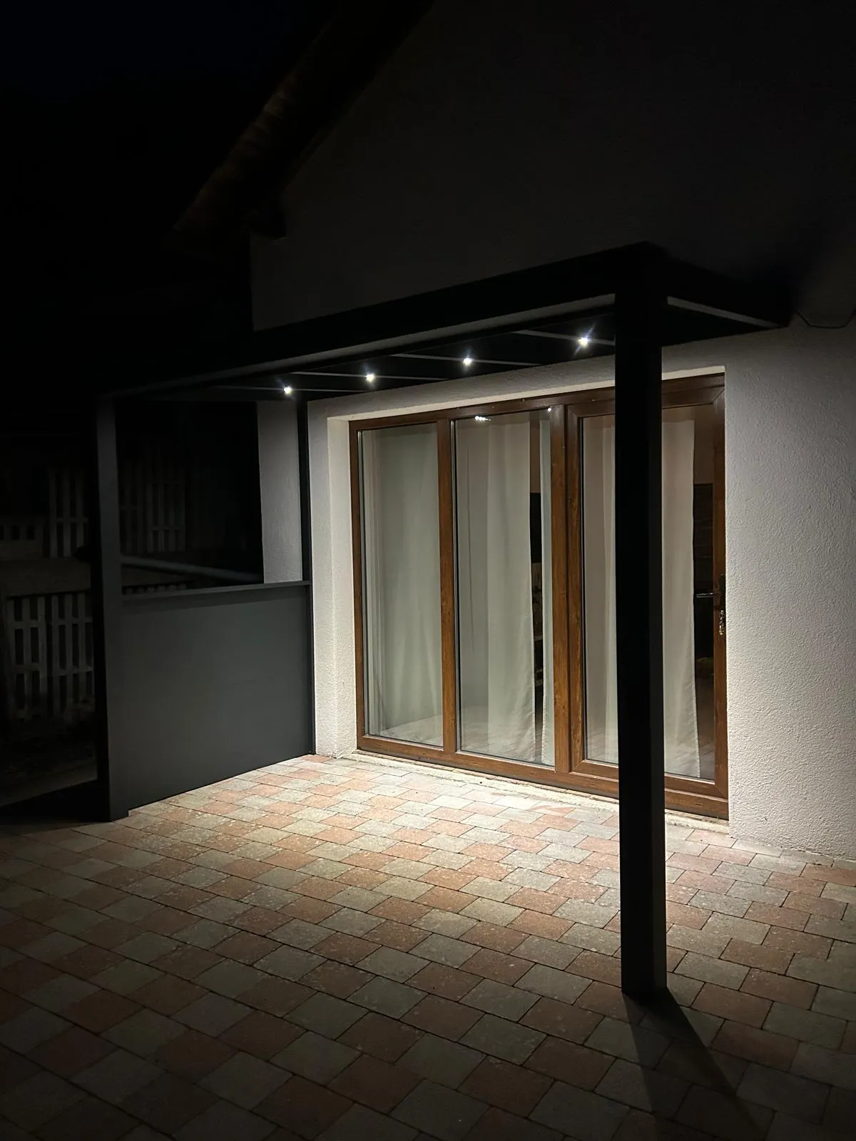 Terrassenüberdachung mit beleuchtetem Pflaster und seitlichem Sichtschutz bei Nacht.